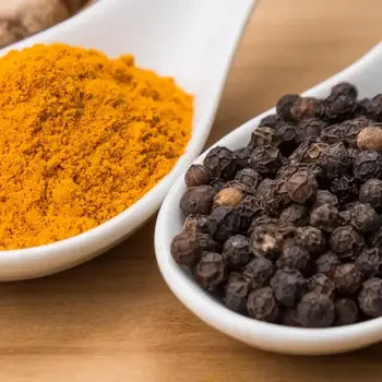 Deux épices dans des cuillères blanches : du curcuma en poudre jaune-orangé et des grains de poivre noir.