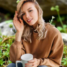 Une femme souriante en plein air, tenant une tasse, entourée de verdure et de fleurs.
