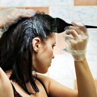 L’image montre une femme appliquant de la teinture sur ses cheveux à l’aide d’un pinceau, tout en portant des gants en plastique.