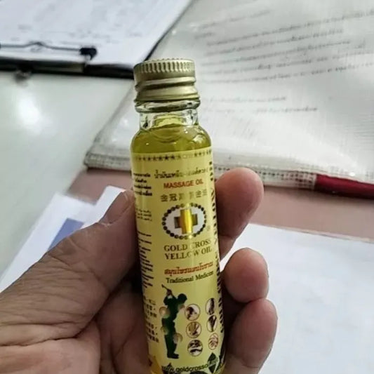 Main tenant une petite bouteille de Gold Cross Yellow Oil, huile traditionnelle thaïlandaise utilisée pour apaiser rapidement douleurs musculaires, tensions et inconforts. Étiquette jaune avec croix dorée et pictogrammes d’utilisation.