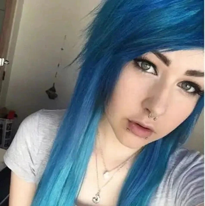Persona con cabello azul vibrante, que lleva un collar y una camiseta gris, fondo borroso con una puerta y decoraciones de pared.