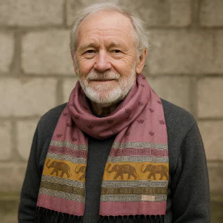 Homme âgé souriant, pull gris et écharpe rose à motifs d’éléphants, devant mur de pierre.