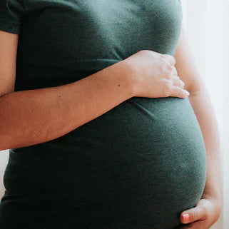 Femme enceinte tenant son ventre, vêtue d’un haut vert, lumière naturelle en arrière-plan.