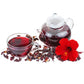 Tasse et théière en verre transparent remplies d'une infusion rouge rubis de fleurs d'hibiscus séchées, éparpillées autour des récipients, avec une fleur d'hibiscus rouge fraîche posée sur fond blanc.