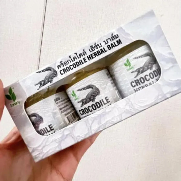 Boîte contenant trois pots de Crocodile Herbal Balm tenue en main. Baume traditionnel utilisé pour apaiser irritations, douleurs légères et inconforts cutanés. Packaging blanc texturé façon peau de crocodile avec illustration de crocodile et feuilles vertes, évoquant un soin naturel et protecteur.