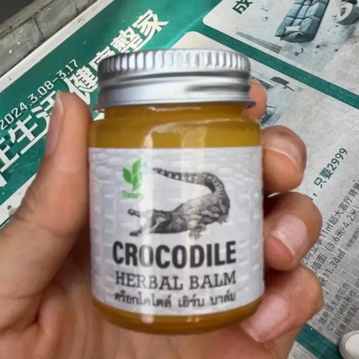 Petit pot de Crocodile Herbal Balm tenu en main, contenant un baume jaune. Baume traditionnel utilisé pour apaiser irritations, douleurs légères et inconforts cutanés. Étiquette avec crocodile et texte thaï, évoquant un soin puissant et protecteur.