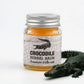 Petit pot de Crocodile Herbal Balm posé à côté d’un crocodile décoratif. Baume à base d’herbes utilisé pour apaiser vite irritations, petites blessures et inconforts cutanés. Packaging avec illustration de crocodile, évoquant un soin puissant et protecteur.