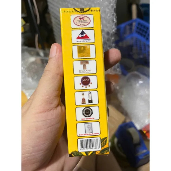 Image d’une boîte jaune présentant plusieurs logos de certifications thaïlandaises, dont des labels de qualité, des marques officielles, des distinctions OTOP et des trophées. La main tient le produit pour montrer ses garanties d’authenticité et de conformité.