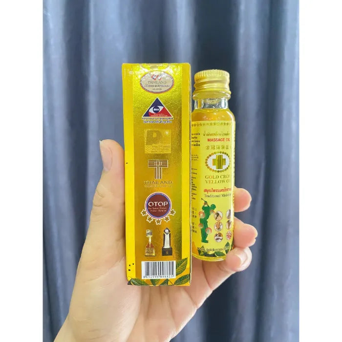 Main tenant une bouteille et sa boîte de Gold Crown Yellow Oil, huile traditionnelle thaïlandaise utilisée pour apaiser rapidement douleurs musculaires, tensions et inconforts. Packaging jaune avec logos de certifications et indications d’usage.